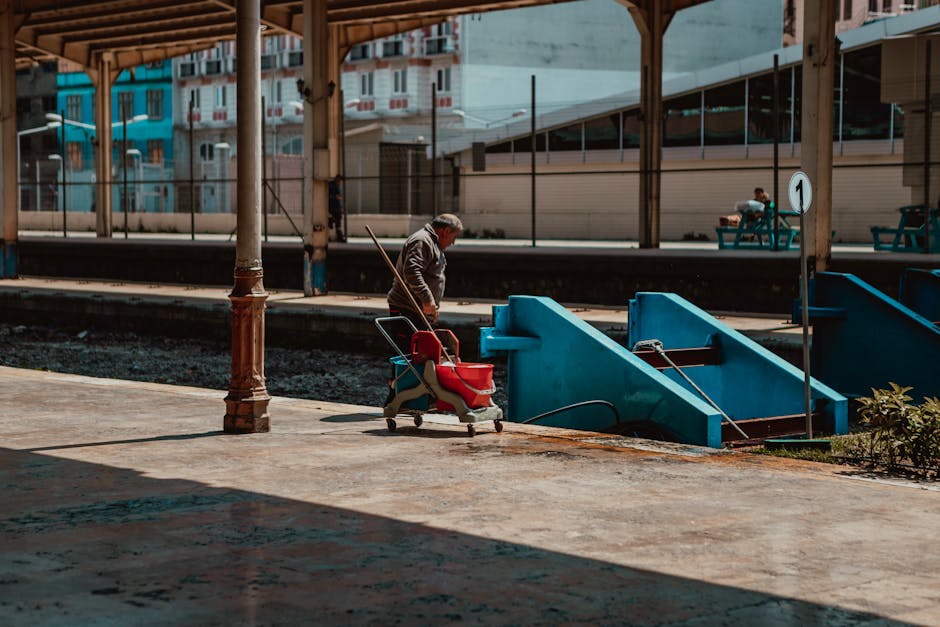 A person with grey hair wearing a grey jacket and dark trousers is walking along a train platform near the blue stairs leading down to the train tracks. They are pushing a small red and grey trolley, 