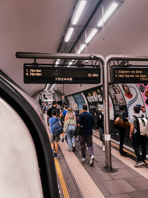 A person with grey hair wearing a grey jacket and dark trousers is walking along a train platform near the blue stairs leading down to the train tracks. They are pushing a small red and grey trolley, 