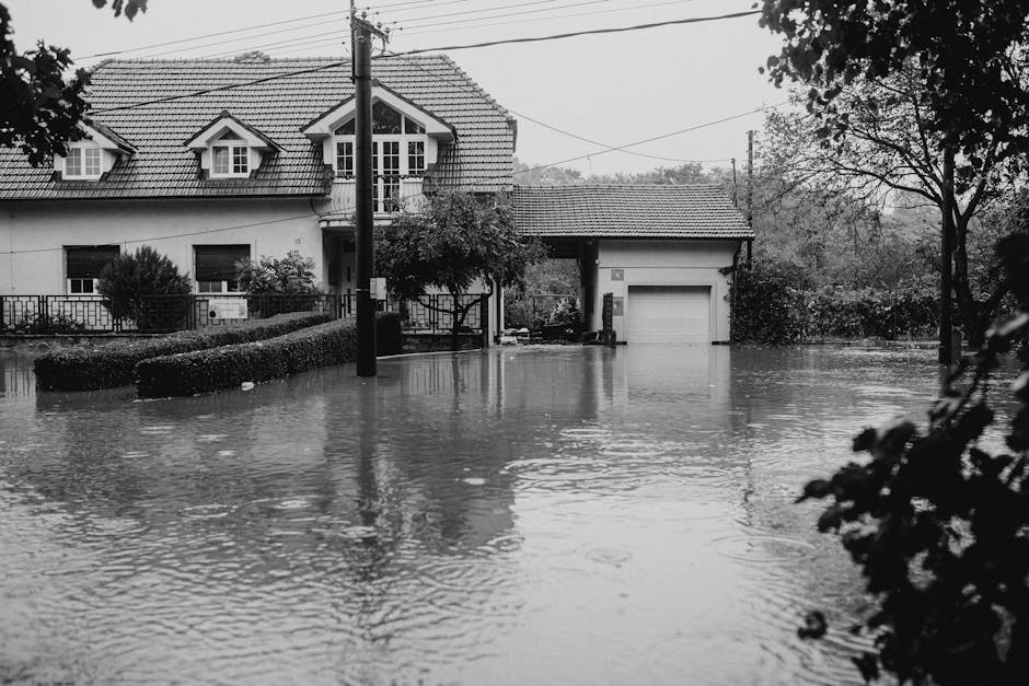 An exterior view of a residential property during a flood, with water covering the front yard, driveway, and partially submerging the garage door. The house has a pitched tiled roof, several small dor